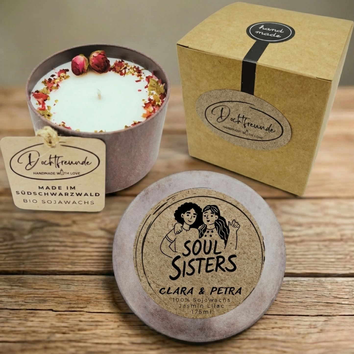 Handgemachte, personalisierte 'Soul Sisters' Sojawachskerze von Dochtfreunde. Das Bild zeigt die offene Kerze mit Blüten- und Rosendekor im braunen Gefäß, den Deckel mit der Aufschrift 'SOUL SISTERS' und den personalisierten Namen 'CLARA & PETRA' sowie die passende Geschenkverpackung aus braunem Karton